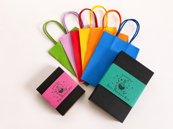 Two Moonatics Eco Potion Kits beside a selection of colourful paper party bags in blue, orange, yellow, red, pink and green, showing a plastic-free party bag alternative for children's celebrations.