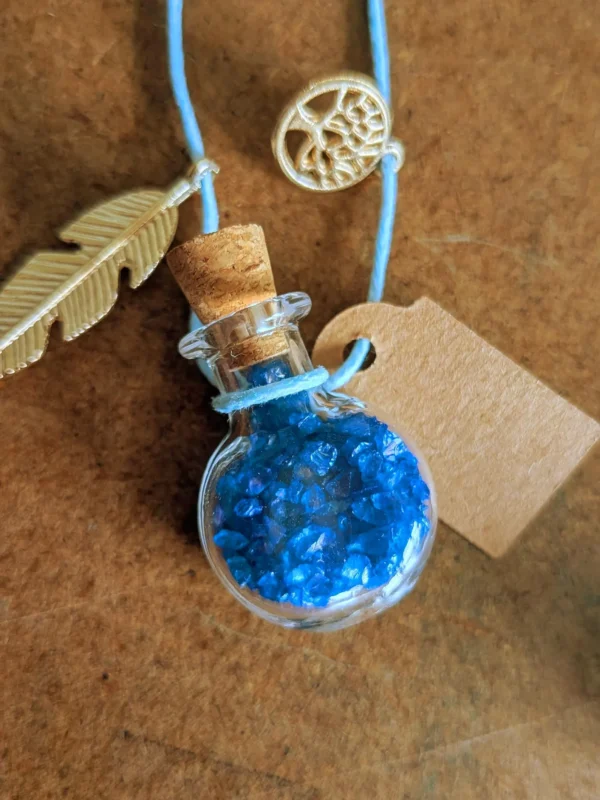 necklace_bottle_round Handmade potion bottle necklace filled with blue crystal gems. The glass bottle hangs from a blue cord and features a cork stopper, metal feather charm, tree charm and blank wish tag.