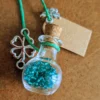 necklace_bottle_sphere Glass potion bottle necklace filled with blue crystal gems, hanging from a turquoise cord with a four-leaf clover charm, cork stopper and blank wish tag.