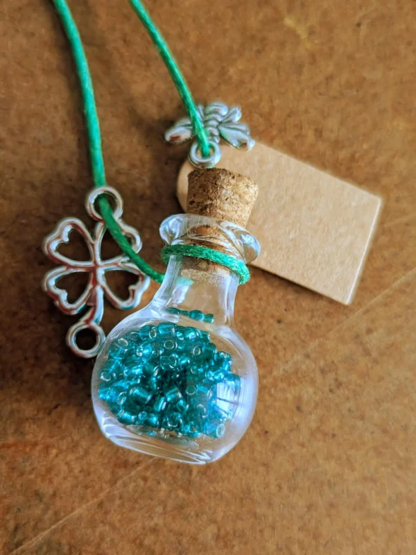 necklace_bottle_sphere Glass potion bottle necklace filled with blue crystal gems, hanging from a turquoise cord with a four-leaf clover charm, cork stopper and blank wish tag.