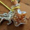 necklace_bottle_star Star-shaped glass potion bottle necklace filled with golden crystal gems. The bottle hangs from a yellow cord and features a cork stopper, unicorn charm and blank wish tag.