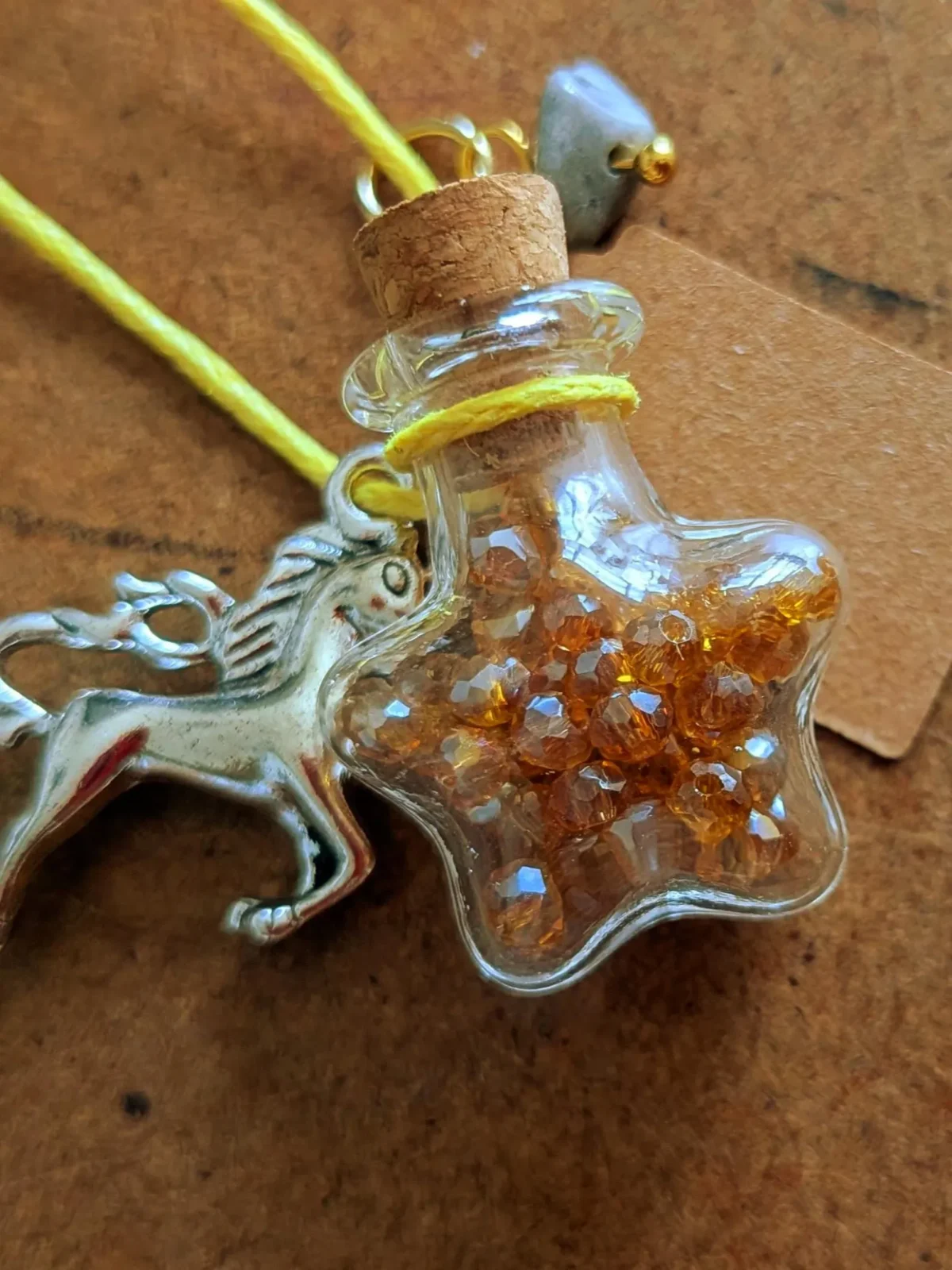 Star-shaped glass potion bottle necklace filled with golden crystal gems. The bottle hangs from a yellow cord and features a cork stopper, unicorn charm and blank wish tag.