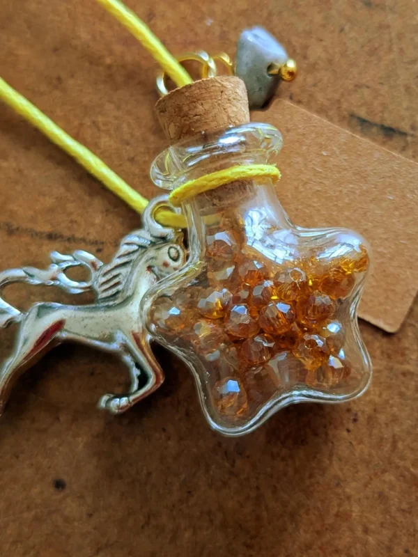 necklace_bottle_star Star-shaped glass potion bottle necklace filled with golden crystal gems. The bottle hangs from a yellow cord and features a cork stopper, unicorn charm and blank wish tag.