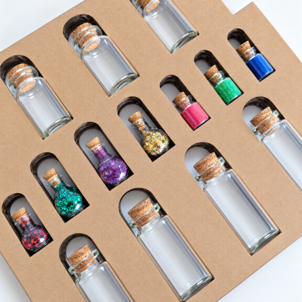 moonatics alchemist eco potion kit - bottles and glitter close up at angle Close-up of glass potion bottles, eco glitter and coloured mica powders arranged in a kraft card tray.